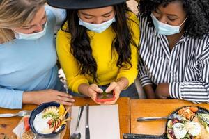 Multiracial friends watching on mobile smartphone while having healthy lunch in coffee brunch bar during corona virus outbreak - Food and technology concept photo