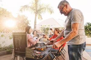 Happy family cheering and toasting with red wine in barbecue party - Chef senior man grilling meat and having fun with parents - Weekend food bbq and reunion young and older people lifestyle concept photo