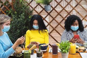 Multiracial friends using mobile smartphone while having healthy lunch in coffee brunch bar during corona virus outbreak - Food and technology concept photo