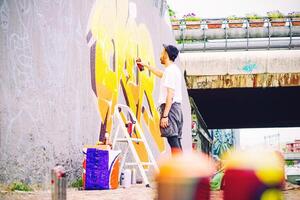 calle artista pintura un vistoso pintada en un gris pared debajo puente - joven grafiter escritura y dibujo murales con amarillo rociar - urbano, estilo de vida y moderno Arte concepto foto