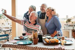 contento personas mayores amigos tomando selfie con móvil teléfono inteligente cámara a cena en terraza - retirado personas teniendo divertido comiendo y Bebiendo rojo vino en patio mientras utilizando nuevo tendencias tecnología teléfono aplicaciones foto