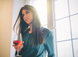 contento joven mujer en pie en su Departamento Bebiendo un vaso de rojo vino siguiente el ventana con un linda sonrisa - hembra disfrutando su hora a hogar con un cóctel - divertido, entretenimiento y estilo de vida concepto foto