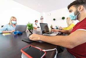 Young people working in modern co-working space wearing surgical mask for preventing corona virus spread - Social distancing, content creator startups and technology concept photo