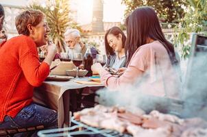 contento familia comiendo y Bebiendo vino a parilla cena en el jardín - personas con diferente siglos comida juntos a barbacoa almuerzo - concepto de comida y bebida foto