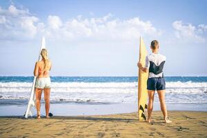 joven Pareja de surfistas en pie en el playa con tablas de surf preparando a navegar en alto olas - sano amigos teniendo divertido con un deportivo día - gente, estilo de vida, deporte concepto foto