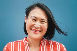 Happy Asian woman portrait against blue background - Senior Chinese female having fun posing in front of camera - Concept of elderly with old generation lifestyle photo