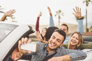 Happy friends taking selfie with mobile smartphone camera in convertible car - Young people having fun in cabriolet auto during road trip vacation - Travel and youth technology lifestyle concept photo