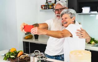 contento mayor Pareja tomando selfie mientras Cocinando juntos a hogar - mayor personas teniendo divertido preparando salud almuerzo en cocina - retirado estilo de vida familia hora y social tecnología concepto foto