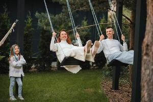 mamá y papá son montando en un columpio y allí es un hija en pie siguiente a a ellos. el familia es descansando en un columpio foto