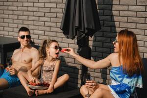 un contento familia en trajes de baño toma el sol en su terraza en verano. mamá alimenta su hija fresas foto