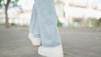 un posterior ver vídeo sigue un mujer pies caminando abajo un calle en un soleado día. un mujer vistiendo zapatillas es caminando en un ciudad acera. un de la persona piernas son ángulo bajo, reflejando el calentar luz de sol. video