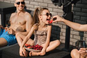 A happy family in swimsuits sunbathes on their terrace in summer. Mom feeds her daughter strawberries photo