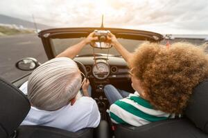 Happy senior couple having fun taking photo with old vintage camera while driving on new convertible car during road trip tour vacation