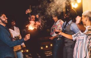 contento joven amigos bailando y celebrando nuevo año víspera con bengalas fuegos artificiales mientras Bebiendo cócteles en patio casa fiesta foto