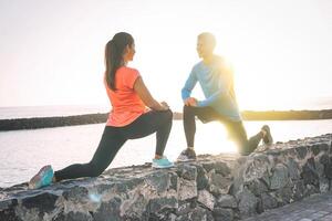 Young health couple stretching legs next to the beach at sunset - Happy sportive lovers workout together standing in front each other and smiling - Relationship, lifetsyle, sport concept photo