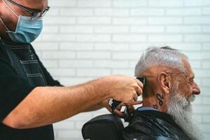 masculino peluquero corte pelo a hipster mayor cliente mientras vistiendo cara quirúrgico máscara - joven estilista trabajando en barbería durante corona virus brote - salud cuidado y Corte de pelo salón concepto foto