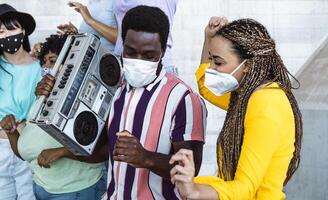 Happy friends listening music with vintage boombox and dancing while wearing face mask outdoor - Multiracial young people having fun during corona virus outbreak - Youth millennial friendship concept photo