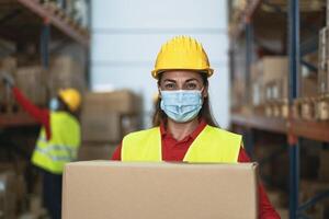 Latin woman working in warehouse carrying delivery boxes while wearing face mask during corona virus pandemic - Logistic and industry concept photo