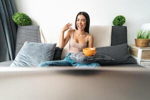 Happy woman eating cereal cup with milk while watching on computer laptop sitting on sofa - Health meal and living room entertainment concept photo