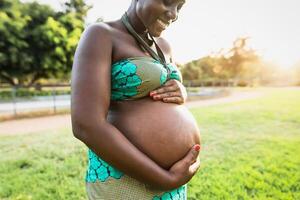 cerca arriba embarazada barriga de joven africano mujer en parque durante puesta de sol hora - maternidad estilo de vida concepto foto