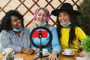 Happy multiracial friends vlogging online using smartphone cam and ring led while drinking a coffee sitting in bar during corona virus outbreak photo