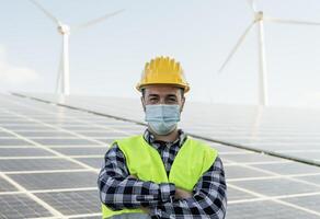 Young engineer working for alternative energy with wind turbine and solar panel while wearing face mask during corona virus outbreak - Innovation and green power concept photo