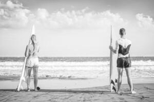 amigos surfistas en pie en el playa con tablas de surf preparando a navegar en alto olas - sano Pareja surf juntos - negro y blanco edición - concepto de deportivo personas estilo de vida foto