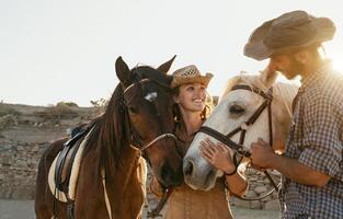 Happy couple having fun with horses inside stable - Young farmers sharing time with animals in corral ranch - Human and animals lifestyle concept photo
