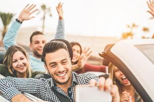 Happy friends taking photo selfie with mobile smart phone camera in convertible car - Young people having fun in cabriolet auto during their road trip vacation - Travel and youth lifestyle concept