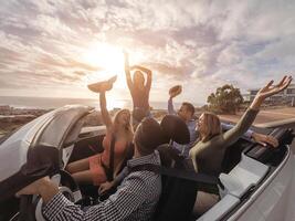 Happy friends with their hands up having fun in cabriolet car on vacation - Young people laughing and enjoying together during travel road trip - Youth holidays lifestyle concept photo