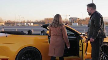 Pareja por amarillo convertible coche a atardecer, un hombre ayuda un mujer obtener dentro un convertible coche. video