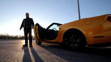 Man Standing Next to a Yellow Convertible Car, Confident man standing next to his yellow convertible car, parked in an open area, with the sun rising in the background. video
