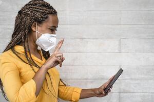 African woman wearing face medical mask using mobile smartphone - Young girl with braids having fun doing video call with phone during corona virus outbreak photo