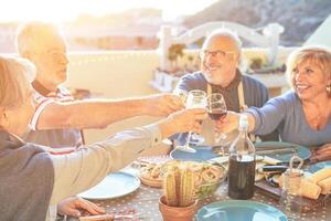 contento mayor amigos teniendo divertido aplausos con rojo vino a parilla en terraza al aire libre - más viejo personas haciendo cena tostado lentes y riendo juntos - amistad y mayor estilo de vida concepto foto