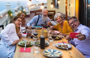 Happy multiracial senior friends having fun dining together while taking selfie with mobile smartphone on house patio - Elderly lifestyle people and food concept photo