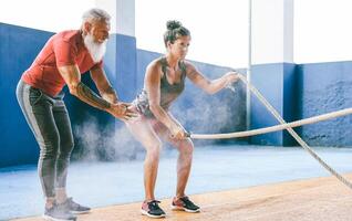 Fit woman training with battle rope inside gym - Hipster personal trainer motivating a female athlete who's doing endurance exercises - Workout and sport activities concept photo