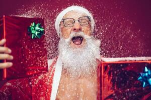Happy crazy santa claus holding christmas box gifts - Hipster sanior man having fun laughing and wearing xmas winter costume - Concept of people doing funny celebration of x-mas holidays photo