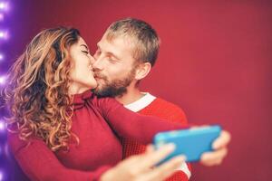 Happy couple taking selfie with mobile smartphone camera - Young romantic lovers kissing and celebrating Christmas holidays - Love relationship, xmas and technology trends concept photo