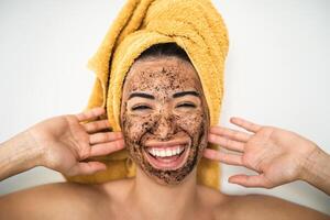 joven sonriente mujer aplicando café fregar máscara en cara - contento niña teniendo piel cuidado spa día a hogar - sano alternativa natural exfoliación tratamiento y juventud personas estilo de vida concepto foto