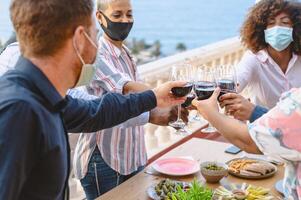 Young multiracial friends toasting with red wine glasses while wearing face protective mask during corona virus pandemic photo