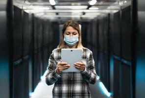 hembra informático ingeniero trabajando dentro servidor habitación base de datos mientras vistiendo cara máscara durante corona virus brote foto