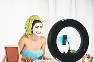 joven hembra vlogger aplicando protección de la piel facial máscara mientras grabación vídeo para Blog a hogar - contento niña teniendo divertido rodaje web tutorial con móvil teléfono inteligente - social influencia estilo de vida concepto foto