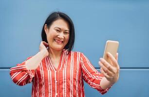 Happy Asian woman using mobile smartphone outdoor - Senior having fun with new trendy technology for social media apps - Elderly lifestyle tech and aged people concept photo