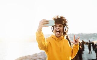 Young Afro man taking self video while listening music with wireless headphones - Happy mixed race guy having fun with new technology apps for mobile smartphone - Millennial lifestyle concept photo