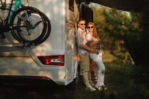 un casado Pareja con lentes soportes siguiente a su casa rodante a puesta de sol en el bosque foto