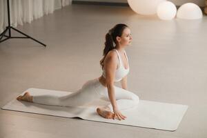 un niña en blanco ropa lo hace yoga en un estera adentro foto