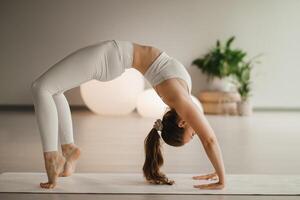 A girl in white clothes does yoga standing on the bridge on a mat indoors photo