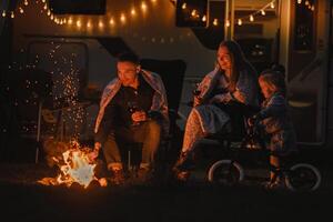 el familia es descansando en el noche cerca el hoguera y su casa rodante foto