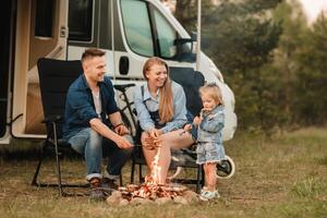 un familia cocineros salchichas en un hoguera cerca su casa rodante en el bosque foto