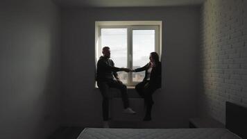 Silhouette of a couple reaching out their hands to each other while sitting by the window, A couple holding hands in a minimalist bedroom with a large window, silhouetted against a cityscape. video